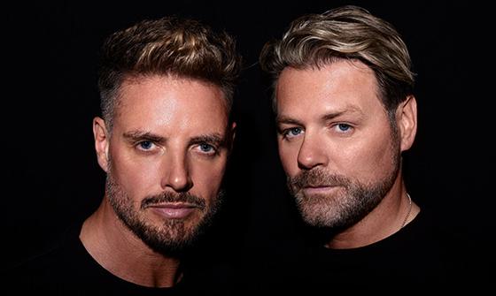 Keith Duffy and Brian McFadden in black tops with a black background looking at the camera
