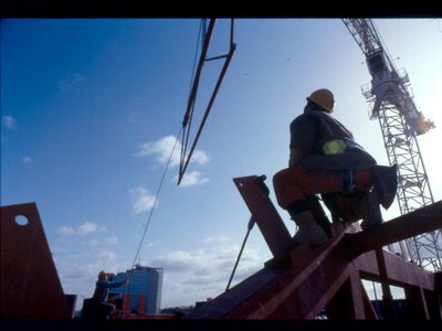 A photo of a builder sat on a beam high above The Anvil watching a crane