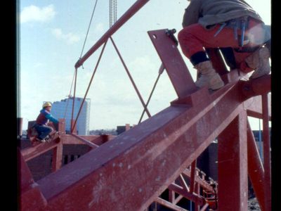 A photo of builder sat high up on a red metal beam as a crane brings round another beam overhead