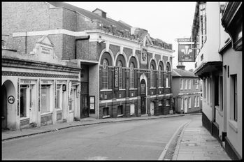 A black and white photo from the top of Wote Street looking down towards the outside of The Haymarket in the 1970s. Notable differences is that there is a small door at the bottom of the arches as well as large doors where the lion wall fountain currently resides. The building now used as the box office and two upper floor foyer is single level and houses two shops called Nicola and F.Scott.