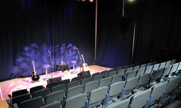A photo taken from the back of The Forge auditorium looking towards the stage which has two stools, guitars and microphones