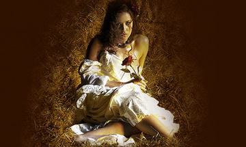 A young woman sits in a pile of straw in a barn with a white prairie dress on holding a red rose.