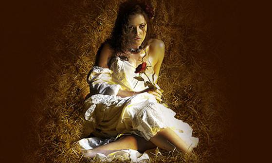 A young woman sits in a pile of straw in a barn with a white prairie dress on holding a red rose