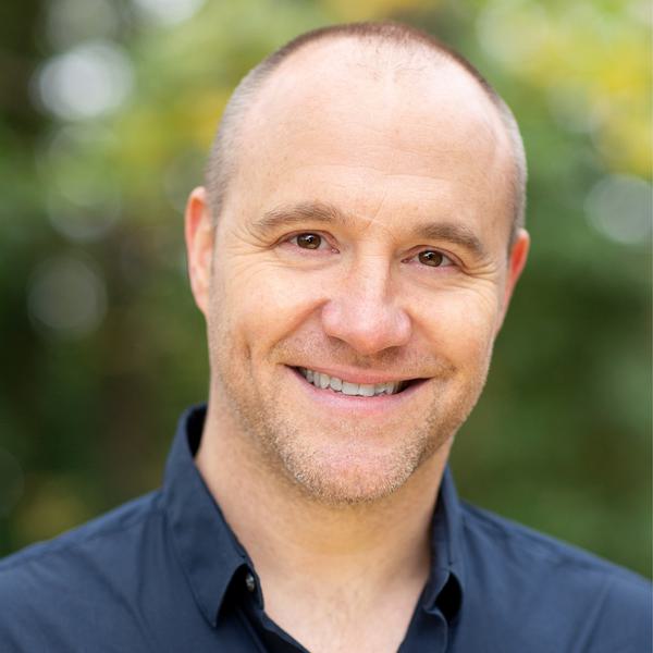 Chris Pizzey a balding white man with a stubbly beard in a dark blue button up shirt smiling at the camera with a blurred forest background
