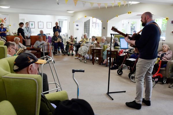 A bald man with a beard playing guitar in front of a room of older people sat in a care home
