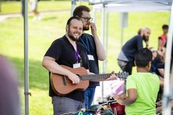 A man holding a guitar and smiling on a field under a gazebo with lots of people around