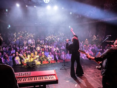 A photo taken from the back of the stage. One guitarist and the keyboard player can both be seen behind the female singer who is performing to the audience