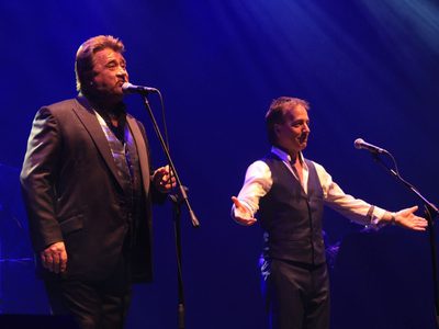 An image to two male singers, David Fawcett and Andy Reiss, performing on stage in black suits
