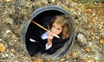 A young girl in a shirt and blazer plays a musical instrument in the woods, she is partially hidden in a submerged pipe