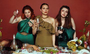 three women stood behind a table holding wine glasses. A fourth woman is passed out on the table