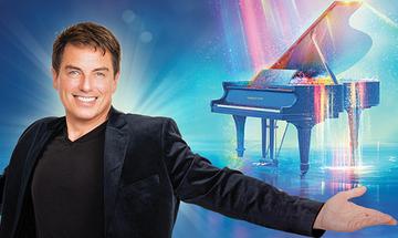 John Barrowman grinning in a black suit holding his arm out presenting a glowing blue piano. All on a blue background with sparkling shafts of a pink and gold lighting