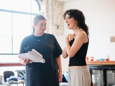 Katy Slater and Kathleen McAuley Irvine rehearse a scene. Katy holds a script and looks at Kathleen, who has a shocked expression and holds her arms crossed over her chest.