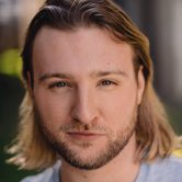 A young white man with a short beard and long dark blonde hair wearing a grey t-shirt