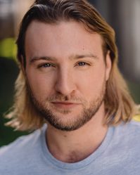 A young white man with a short beard and long dark blonde hair wearing a grey t-shirt