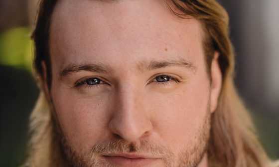 A young white man with a short beard and long dark blonde hair wearing a grey t-shirt