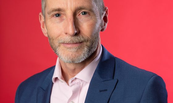 An older man with grey hair stands against a red background, he wears a pink shirt and a navy blazer