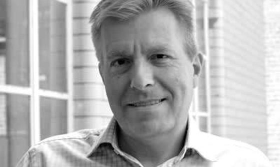 A black and white photo of an older man wearing a checked shirt, stood outside a building