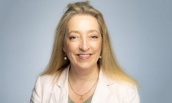 A blonde woman wearing a green top and white blazer smiles