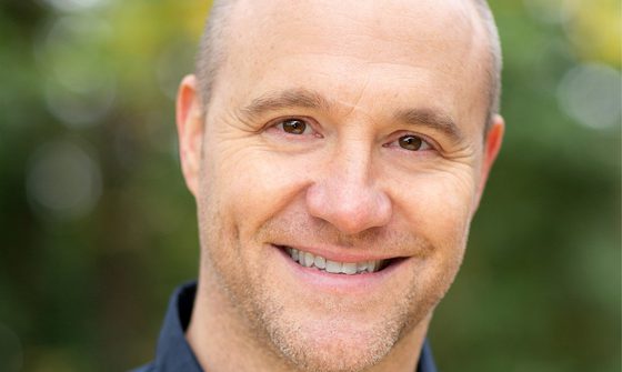 Chris Pizzey a balding white man with a stubbly beard in a dark blue button up shirt smiling at the camera with a blurred forest background