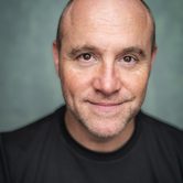 An older bald white man in a black t-shirt with a calm smile
