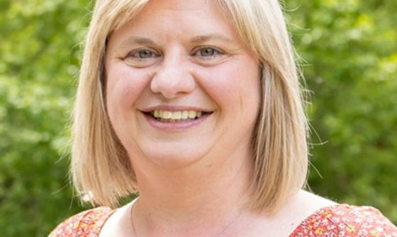 A blonde woman smiles. She's wearing a silver necklace and an orange floral dres