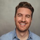 Dan O'Brien, a white man with dark blonde hair styled in a quiff with a stubbly beard and wearing a denim shirt. He is smiling a wide grin at the camera