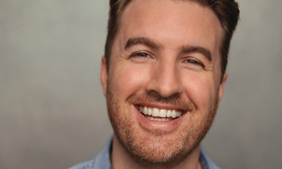 Dan O'Brien, a white man with dark blonde hair styled in a quiff with a stubbly beard and wearing a denim shirt. He is smiling a wide grin at the camera