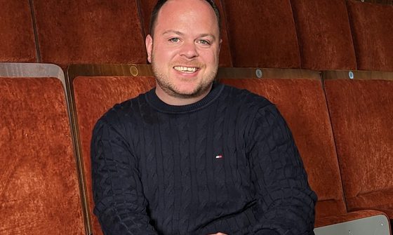 A man with short brown hair, wearing jeans and a navy jumper, sits in a theatre seat with his legs crossed and smiles