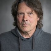 An older man with long greying dark hair in a grey hoodie looking at the camera with a neutral expression