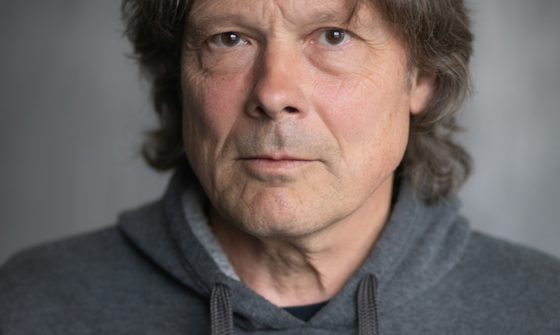 An older man with long greying dark hair in a grey hoodie looking at the camera with a neutral expression