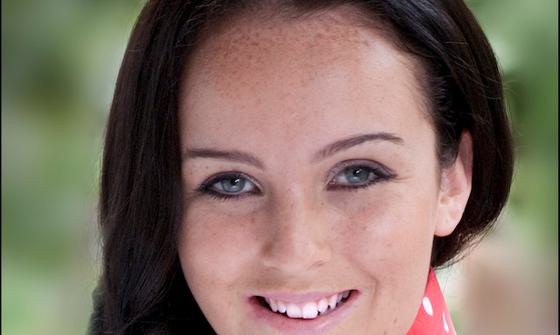 This is a portrait photograph of Juliette Strobel with long, dark brown hair styled in loose waves. She is wearing a sleeveless red blouse with white polka dots. Her expression is cheerful and friendly, with a bright smile.