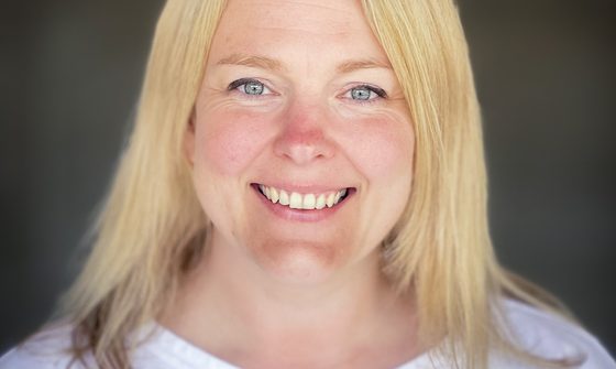 A blonde woman, wearing a white t-shirt, smiles at the camera against a dark green background