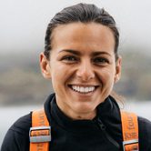 A young white woman with dark hair that is tied back smiling in a black fleece with orange straps in front of a large body of water