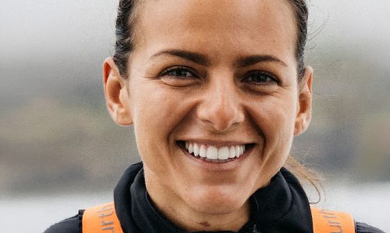 A young white woman with dark hair that is tied back smiling in a black fleece with orange straps in front of a large body of water