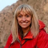 An older white woman with dyed blonde hair in a red coat smiling in front of a mountain