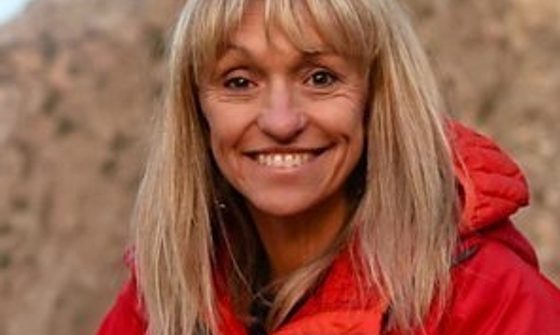 An older white woman with dyed blonde hair in a red coat smiling in front of a mountain