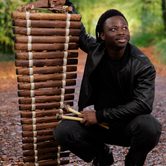 N’famady Kouyaté, a young black man with black hair in a black leather jacket, black top, and black trousers squatting down next to his balafon, and holding the beaters in his hand, in the middle of the woods