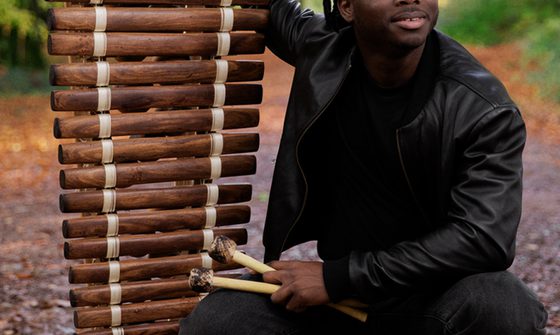 N’famady Kouyaté, a young black man with black hair in a black leather jacket, black top, and black trousers squatting down next to his balafon, and holding the beaters in his hand, in the middle of the woods