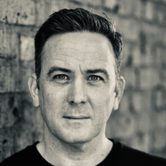 a black and white headshot of Nic Gibney. Nic is wearing a black long sleeved t-shirt and is stood against a brick wall