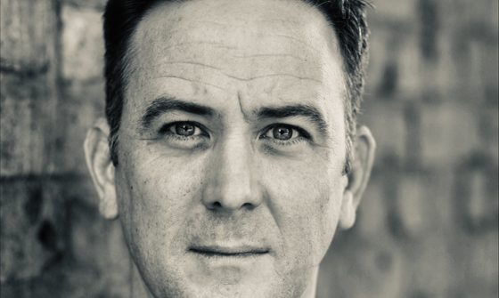 a black and white headshot of Nic Gibney. Nic is wearing a black long sleeved t-shirt and is stood against a brick wall