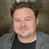 A young white man with quiffed dark brown hair and very short beard wearing a black t-shirt and grey shirt