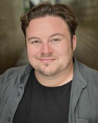 A young white man with quiffed dark brown hair and very short beard wearing a black t-shirt and grey shirt