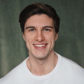 A young white man with quiffed dark brown hair grinning at the camera with a white t-shirt on