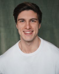 A young white man with quiffed dark brown hair grinning at the camera with a white t-shirt on
