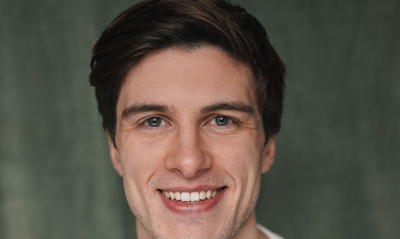 A young white man with quiffed dark brown hair grinning at the camera with a white t-shirt on