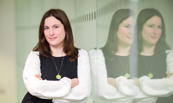 Sara wears a white shirt under a black dress. She is smiling into the camera and has her arms folded