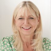 Sarah Jane Buckley, an older white woman with light blonde hair smiling at the camera in a green top with white flowers