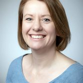 A pale white woman with a brunette bob wearing a light blue top grinning at the camera