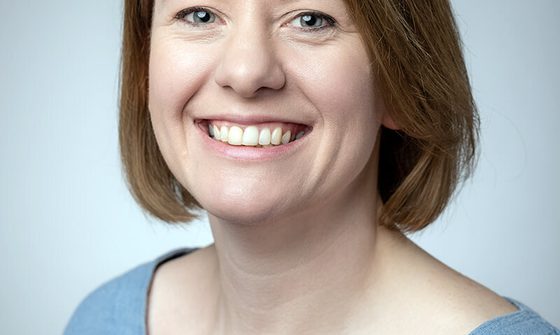 A pale white woman with a brunette bob wearing a light blue top grinning at the camera