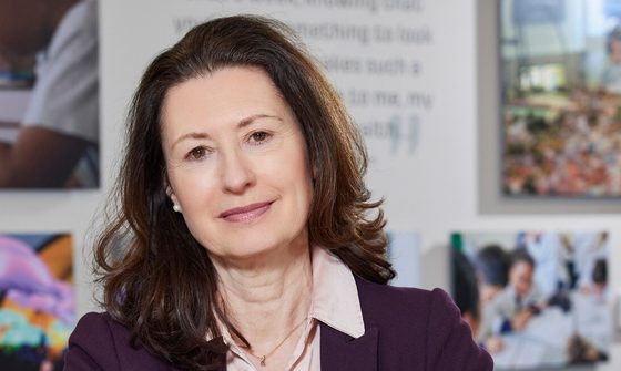 A brunette woman sits at a desk with her arms resting on the table. She's wearing a pink shirt and a purple blazer.